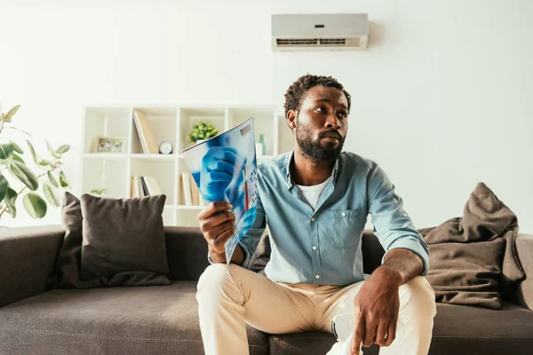 Unhappy African Man Waving Magazine While Sitting Couch Suffering Heat — Stock Photo, Image