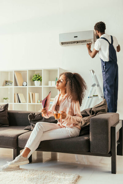 attractive african american woman sitting on sofa with cup of tea, while african american handyman repairing air conditioner