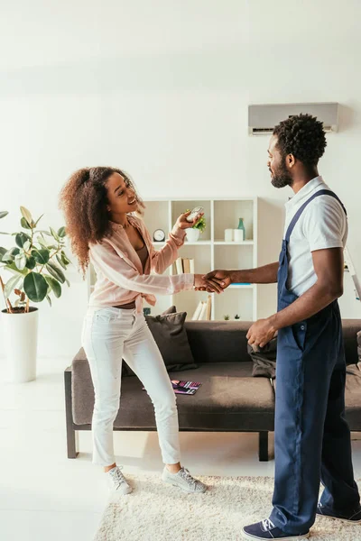 happy african american woman holding air conditioner remote controller while shaking hands with african american repairman