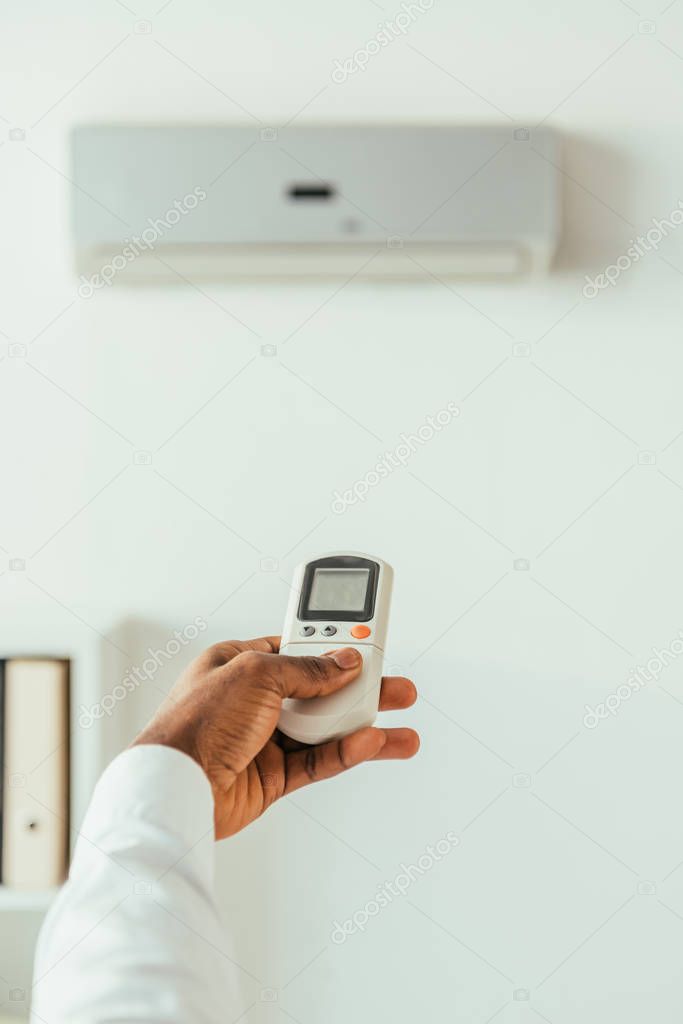 cropped view of african american businessman holding remote controller while standing under air conditioner in office