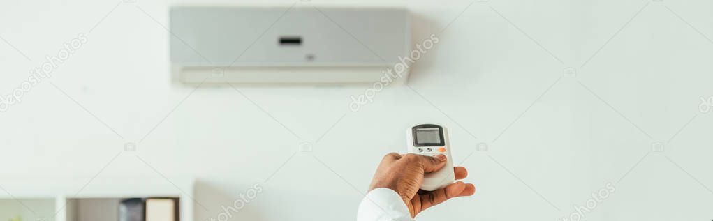 partial view of african american businessman holding remote controller while standing under air conditioner, panoramic shot