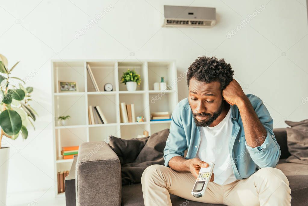 dissatisfied african american man looking at air conditioner remote controller while suffering from heat at home