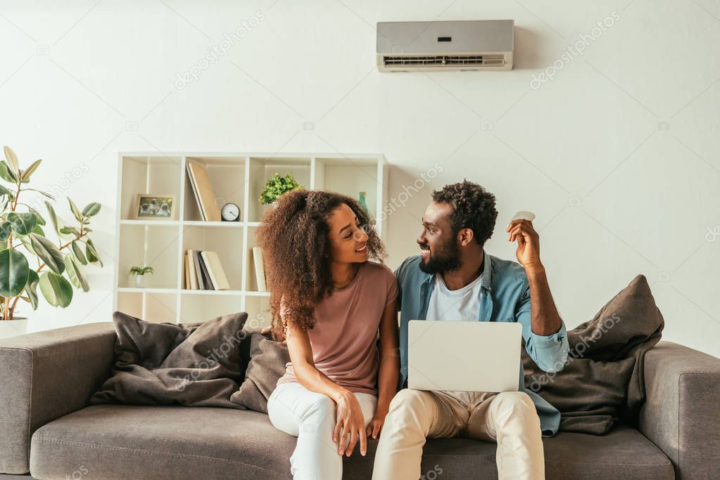 handsome african american man using laptop and holding air conditioner remote controller while sitting on sofa near cheerful african american woman