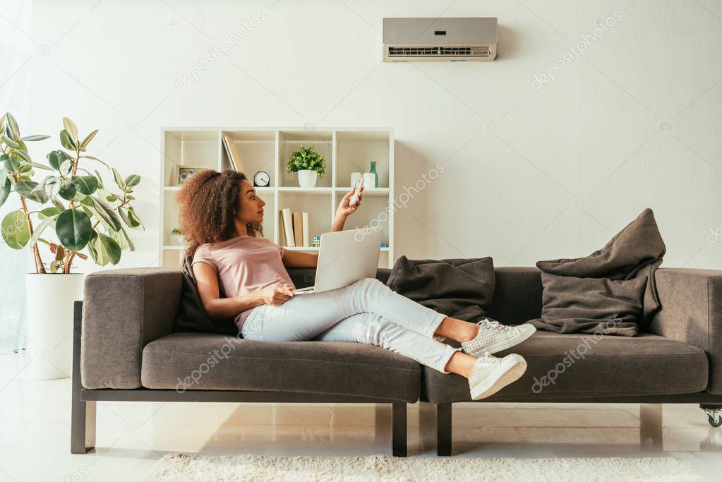 pretty african american woman using laptop and holding remote controller while lying on sofa under air conditioner