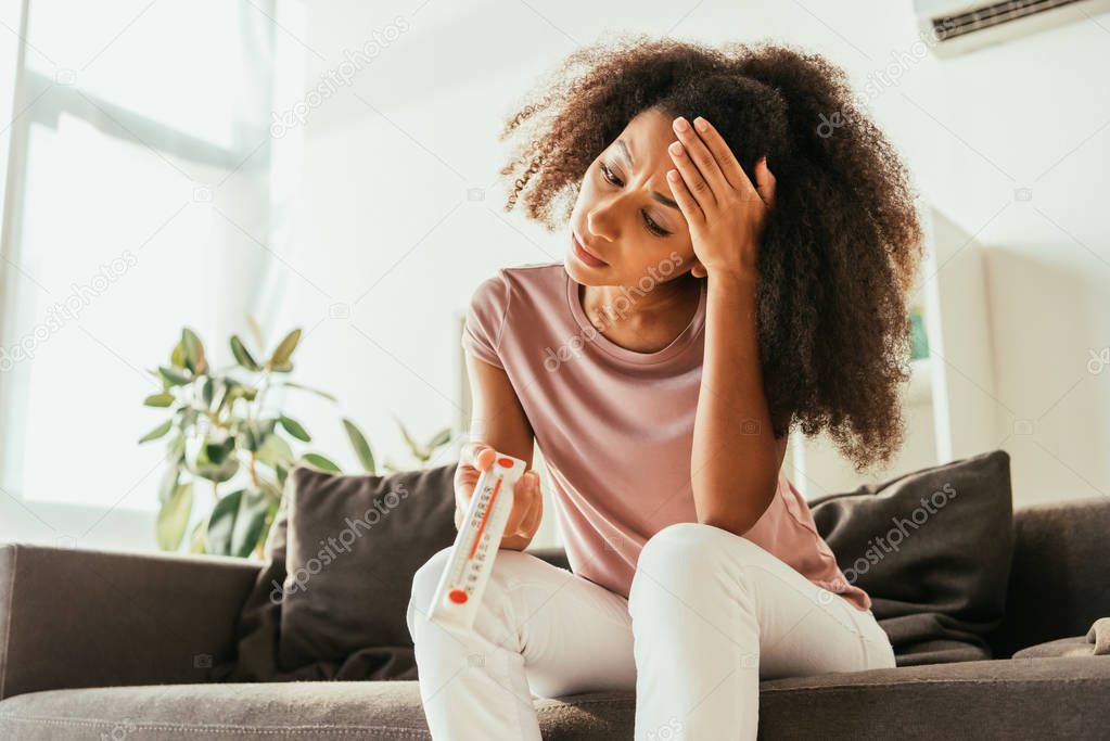 unhappy african american woman looking at thermometer showing high temperature and holding hand on head