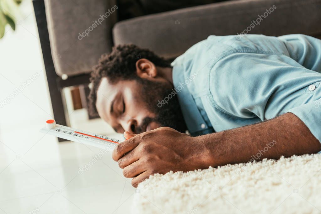 exhausted african american man suffering from summer heat while lying on floor with thermometer in hand