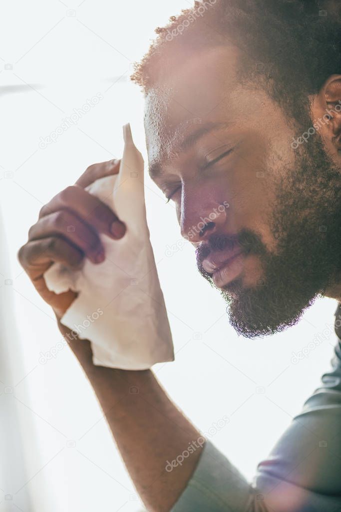 exhausted african american man holding napkin while suffering from summer heat with closed eyes
