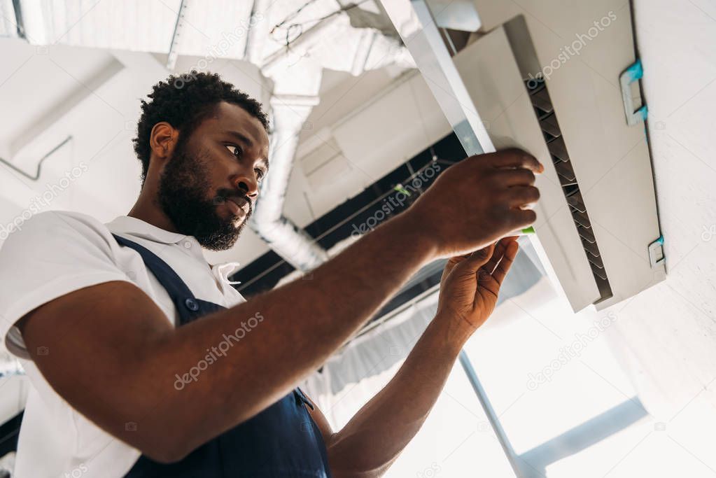 concentrated african american repairman standing on ladder and fixing air conditioner