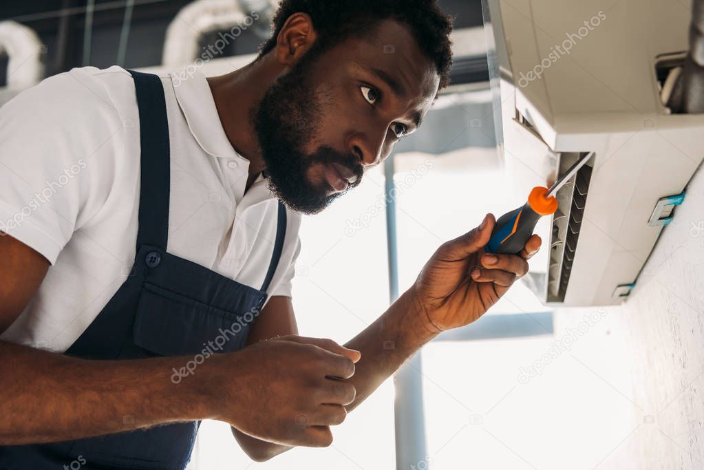 attentive african american repairman fixing air conditioner with screwdriver