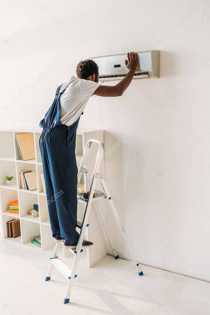 african american handyman in overalls standing on ladder and repairing air conditioner