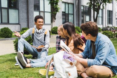 smiling teenagers sitting on grass, talking and holding books  clipart