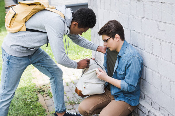 african american boy taking backpack from frightened boy in glasses 