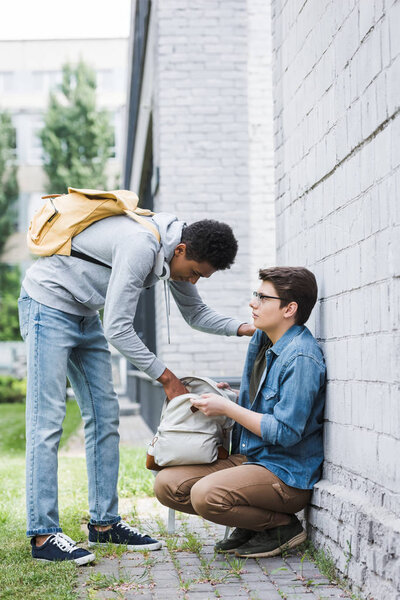 african american boy taking backpack from frightened boy in glasses 
