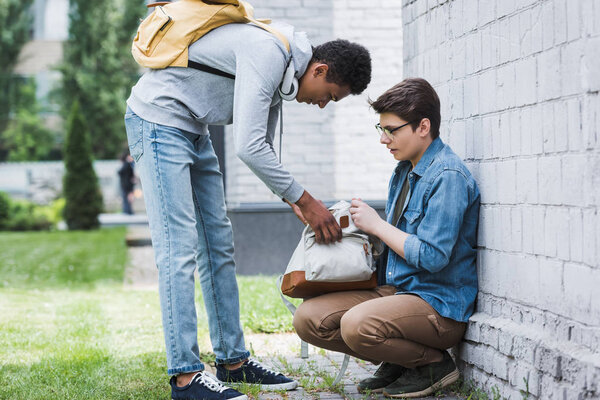 aggressive african american boy taking backpack from frightened boy in glasses 