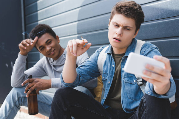 избирательный фокус друзей, улыбающихся, курящих сигареты, держащих пиво и делающих селфи
 