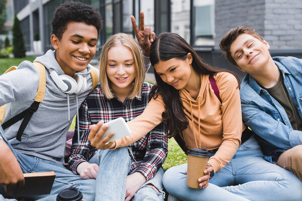 smiling and happy teenagers sitting on grass and taking selfie 