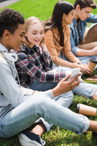 smiling and happy teenagers sitting on grass and looking at smartphone 