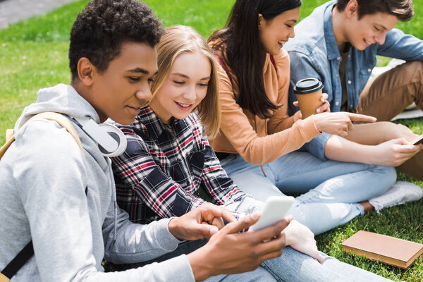 smiling and happy teenagers sitting on grass and looking at smartphone 