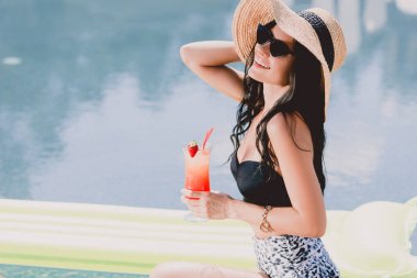 mutlu güzel esmer genç kadın mayo, hasır şapka ve güneş gözlüğü su yakınında kokteyl tutarken gülümseyerek