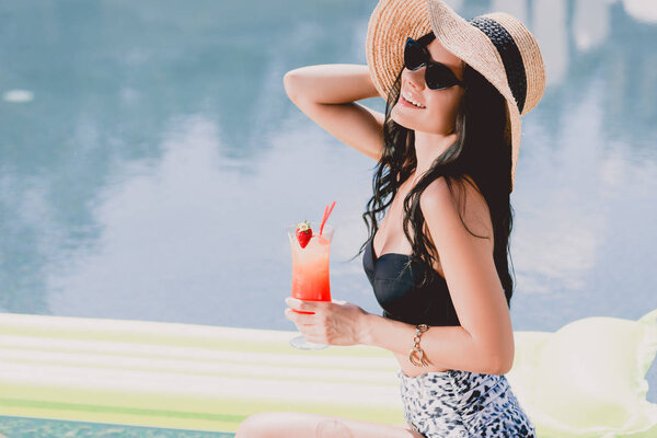 happy beautiful brunette young woman in swimsuit, straw hat and sunglasses smiling while holding cocktail near water