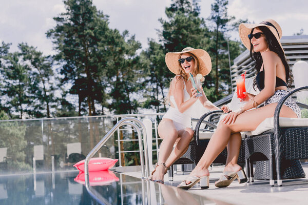 счастливые девушки, сидящие с коктейлями возле бассейна

