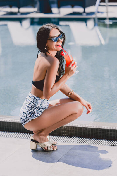 happy sexy brunette woman in swimsuit and sunglasses sitting near swimming pool and drinking cocktail 