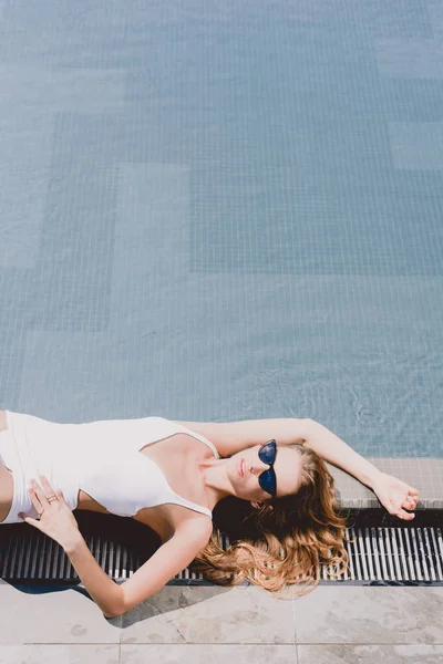性感的金发女郎在太阳镜和白色泳衣躺在游泳池附近 — 图库照片