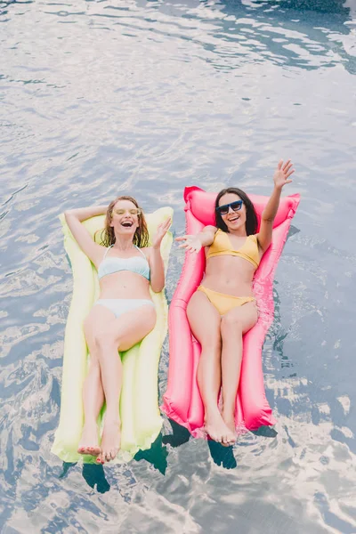 黑发和金发女郎的头顶视图快乐和兴奋的妇女躺在游泳池浮子上伸出双手在游泳池 — 图库照片