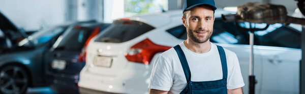 panoramic shot of happy handsome car mechanic looking at camera near cars 