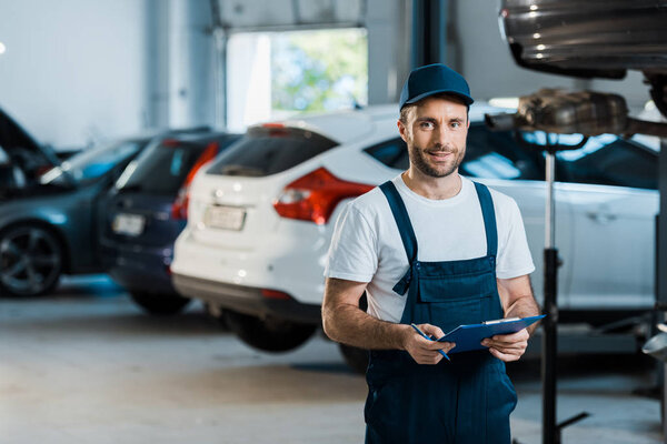 happy bearded car mechanic looking at camera and holding clipboard near cars 