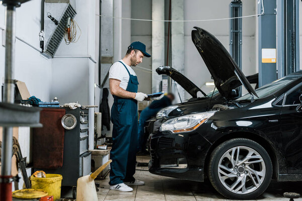 handsome repairman in cap holding pen and clipboard near car
