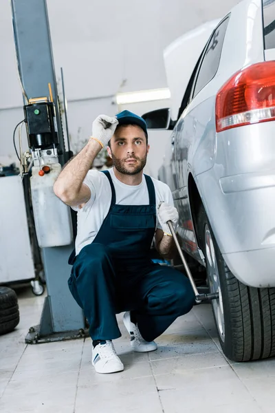 Bearded Car Mechanic Uniform Touching Cap While Changing Car Tire — Stock Photo, Image