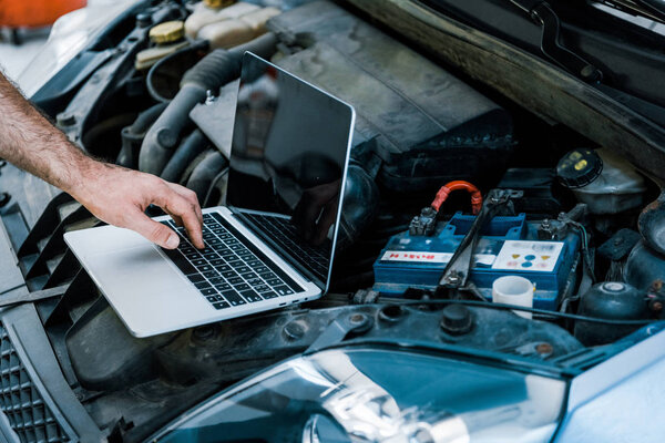 cropped view of car mechanic using laptop with blank screen near car 