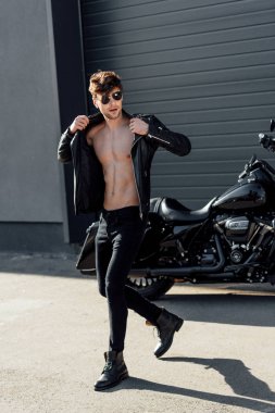 handsome young man taking off leather jacket while walking near motorcycle clipart