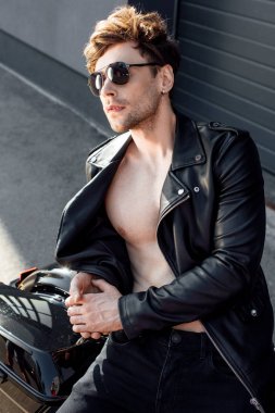 young man with nude chest in leather jacket leaning on motorcycle clipart