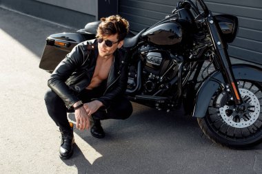 muscular young man sitting near motorcycle and looking away clipart