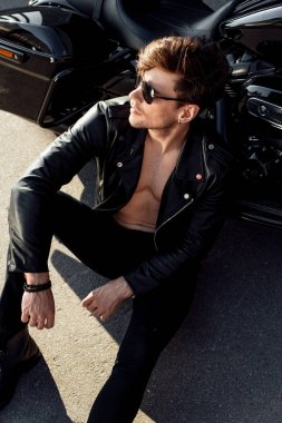 handsome man with muscular torso sitting on ground near motorcycle and looking away clipart