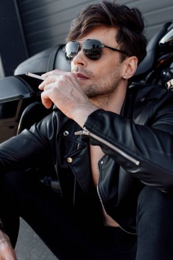 motosiklet yakınında otururken sigara tutan güneş gözlüğü ve deri ceket genç adam