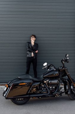 duvara yaslanmış ve motosiklet yakınında duran sigara içen genç adamın tam uzunlukta görünümü