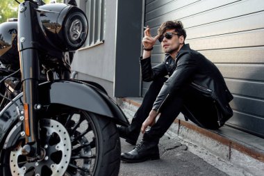 güneş gözlüğü yakışıklı motosiklet yakınında yerde oturan ve sigara tutan yakışıklı motosikletçi 