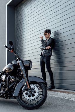 motosiklet yakın duvar ayakın duran ve sigara tutan güneş gözlüğü yakışıklı motosiklettam boy görünümü 