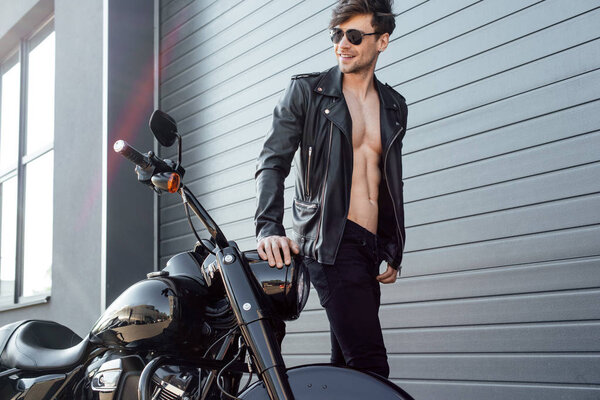  young man in sunglasses with leather jacket and naked torso standing near black motorcycle, smiling and looking away 