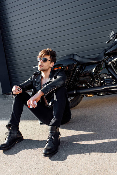 Молодой человек с сигаретой сидит на земле возле черного мотоцикла и держит зажигалку
 