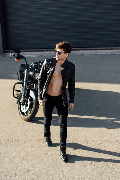 полный вид молодого человека с мускулистым туловищем в кожаной куртке, смотрящего в сторону и курящего сигарету
