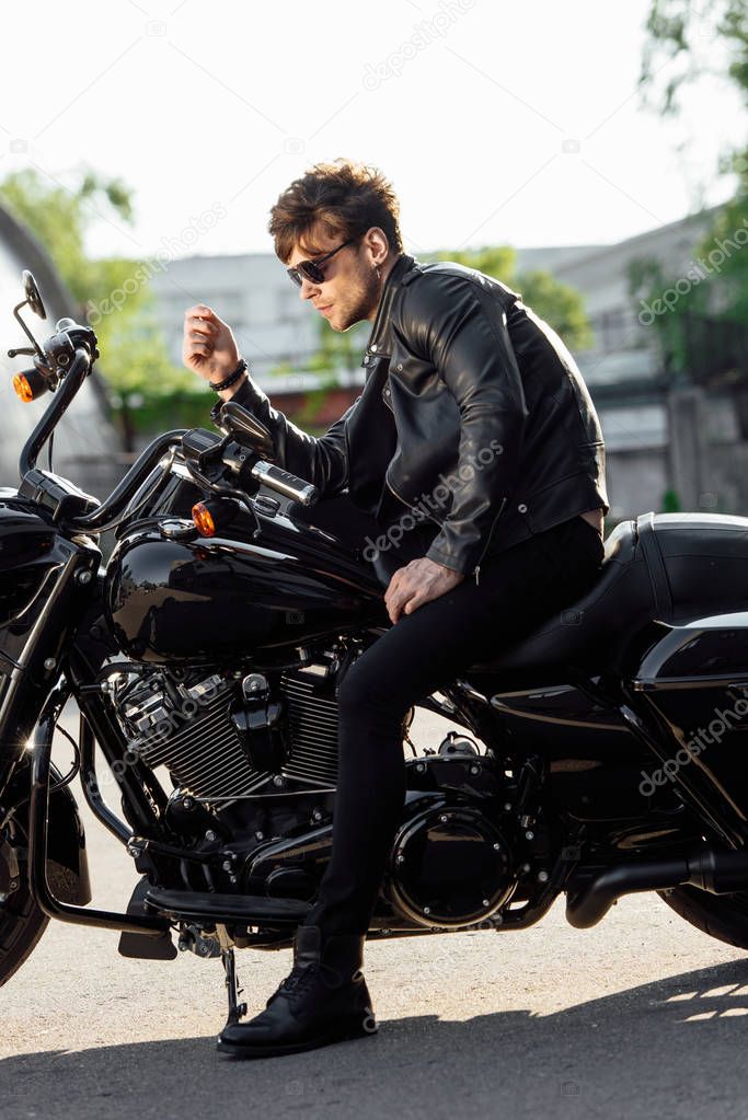 side view of motorcyclist in sunglasses and leather jacket sitting on motorcycle