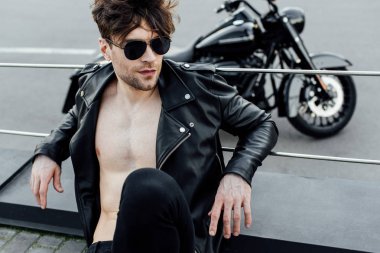 selective focus of man with naked torso in leather jacket sitting on ground with motorcycle on background clipart