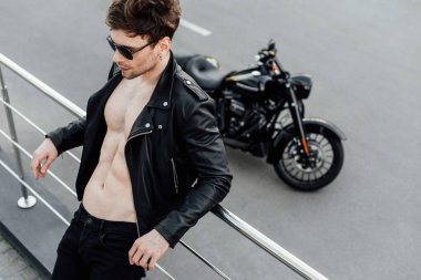 selective focus of man in leather jacket with naked torso leaning on metal fence standing not far from motorcycle clipart