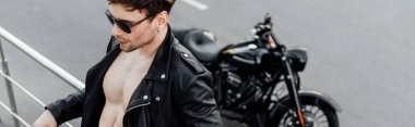 panoramic shot of man in leather jacket with naked torso leaning on metal fence standing not far from motorcycle clipart