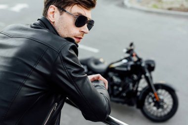 arka planda motosiklet ile metal çit yanında duran siyah deri ceket genç adamın seçici odak