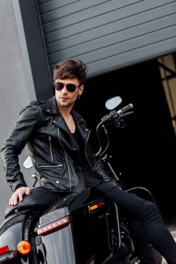 garaj yakınında motosiklet üzerinde oturan güneş gözlüğü ve deri ceket motosikletçi